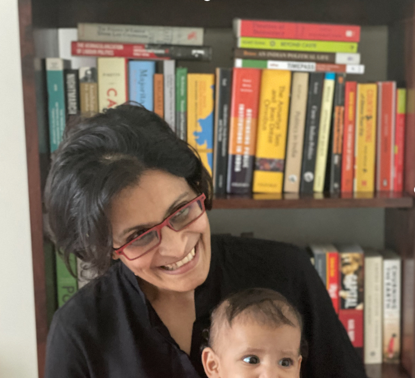 This is an image of Dr. Sai Balakrishnan holding her daughter. She is sitting in front of a bookcase, while smiling.
